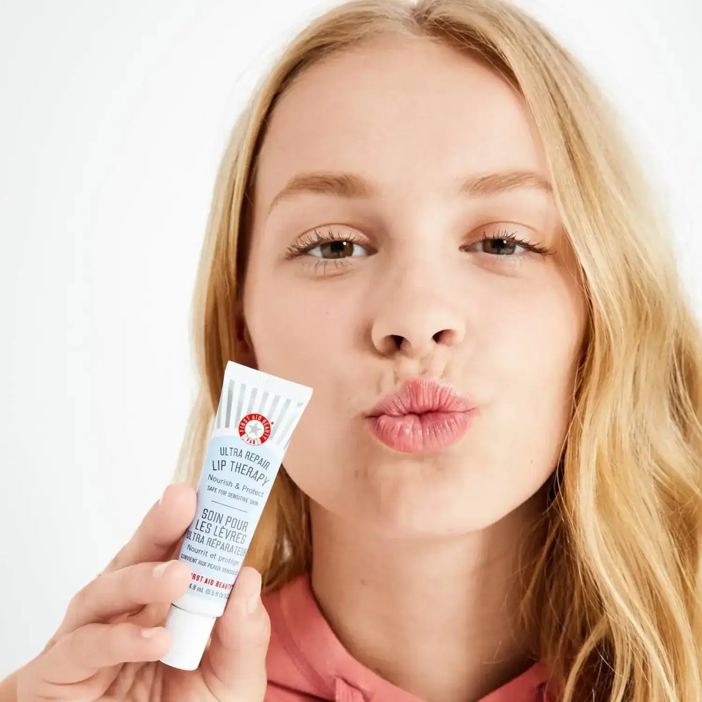 Young woman holding First Aid Beauty Ultra Repair Lip Therapy, showcasing moisturized lips with a kiss pout.