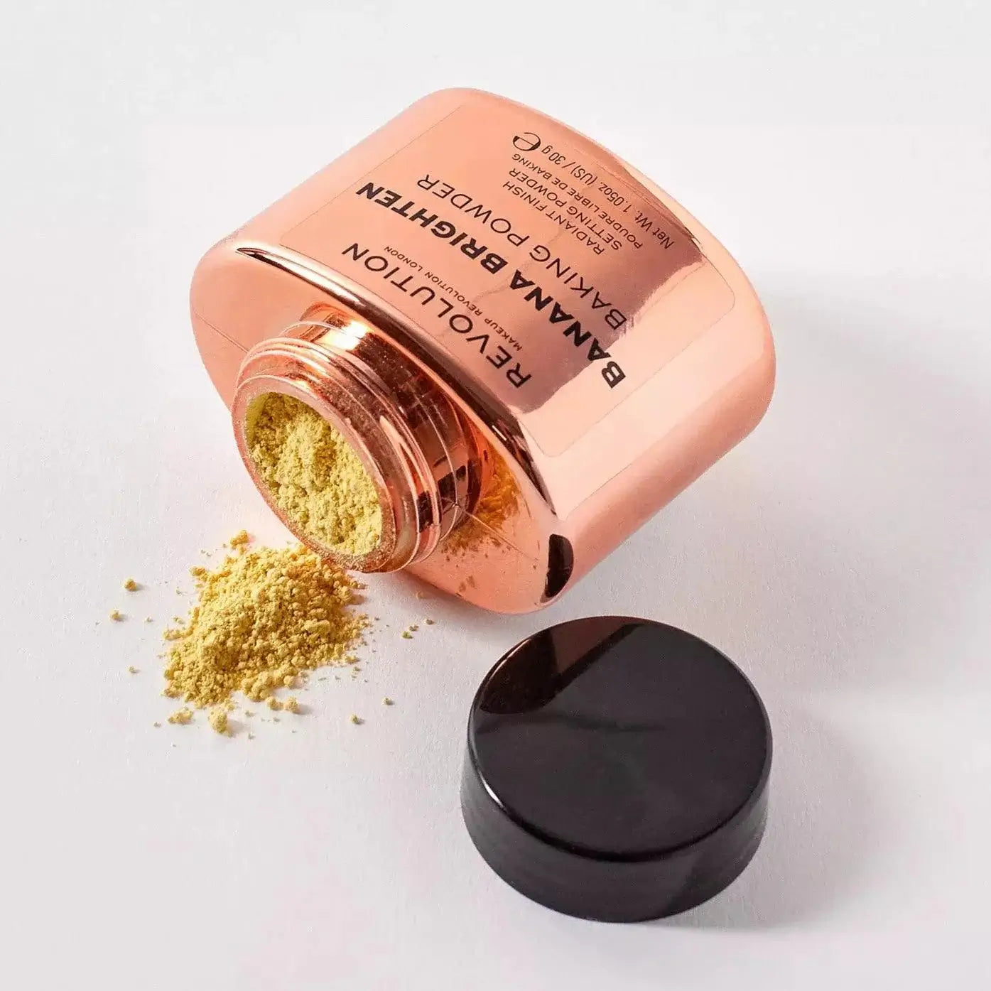 Makeup Revolution Banana Brighten Loose Baking Powder with golden hue spilling from jar on white background.