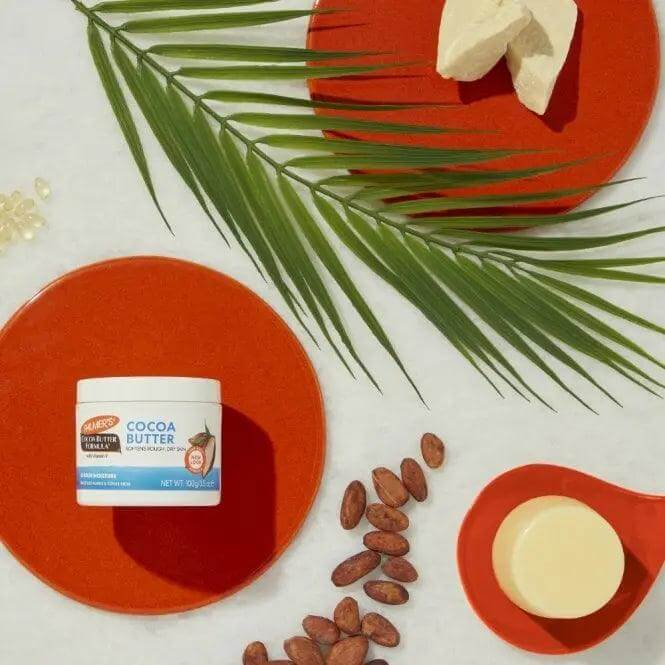 Palmer's Cocoa Butter Original Solid Formula on red plates with cocoa beans and shea butter for skin moisturizing.