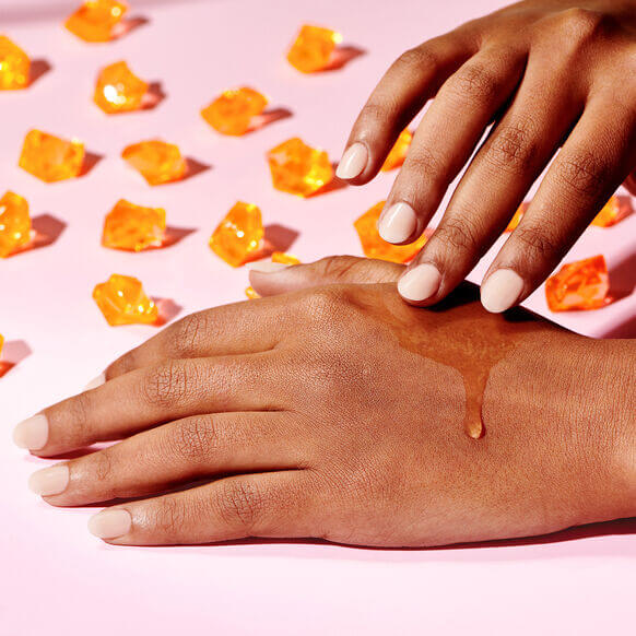 Hand applying Drunk Elephant C-firma Fresh Day Serumon a pink background with orange cosmetic products.