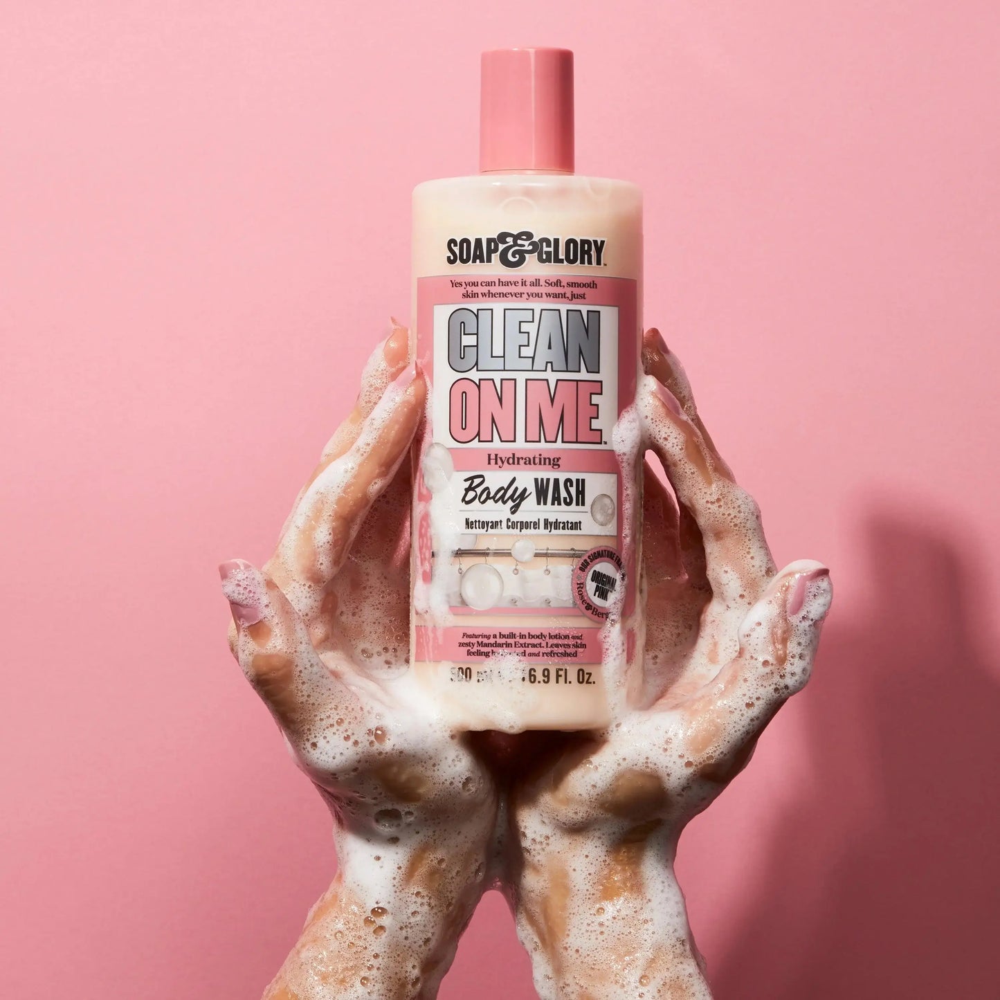 Soap & Glory Clean on Me Body Wash in a bottle held above soapy hands against a pink background.