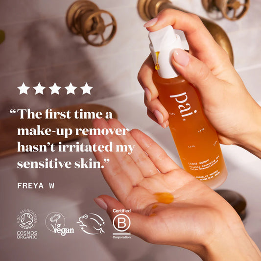 Hand holding a bottle of Pai skincare product with a quote and certification logos on a blurred bathroom background.