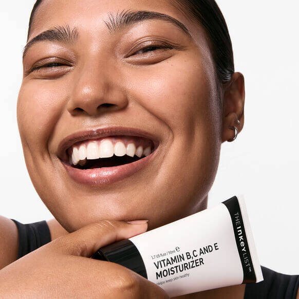 Smiling woman holding The INKEY List Vitamin B, C and E Moisturizer 50ml, showcasing healthy, glowing skin.
