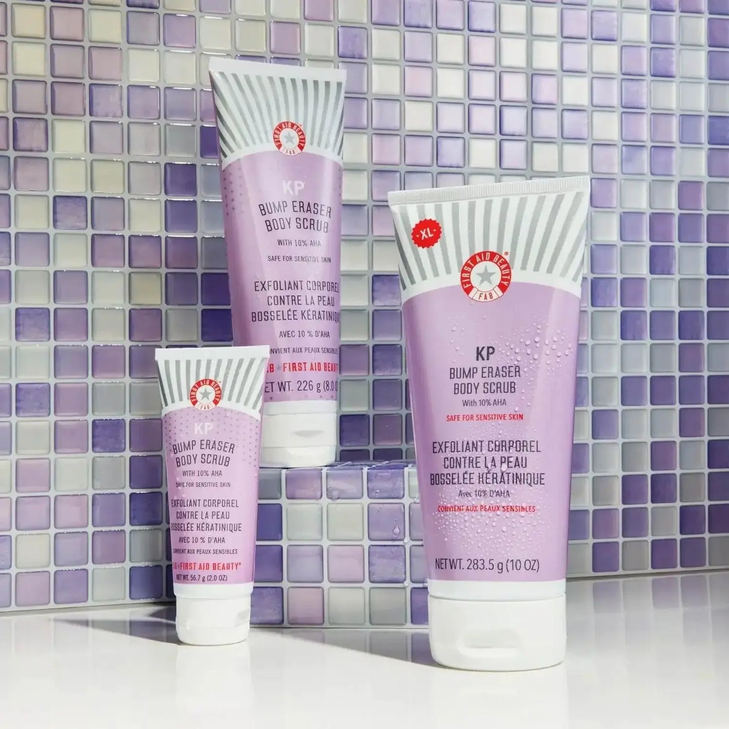 First Aid Beauty KP Bump Eraser Body Scrub in three travel sizes against a purple tiled background.