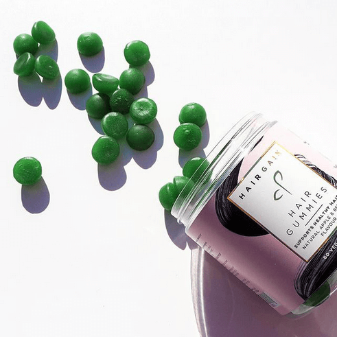 Hair Gain Gummies in a jar with green gummy supplements scattered on a white surface for healthy hair.