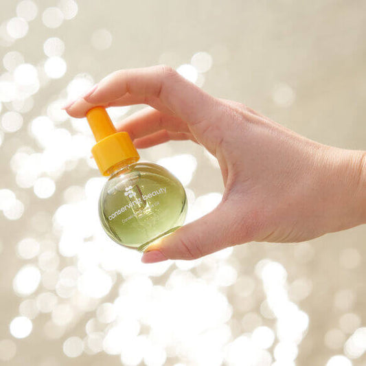 Hand holding Conserving Beauty Conserve You Face Oil bottle with a shimmering background, highlighting its radiant skin benefits.