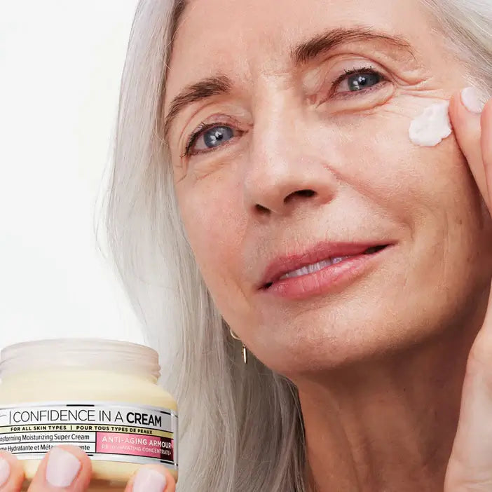 Woman applying IT Cosmetics cream to her face with a jar of 'Confidence in a Cream' in the foreground.