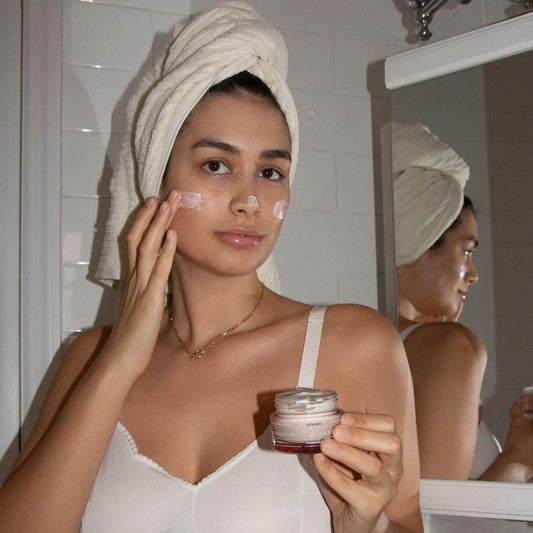 Woman applying Korres cream to her face with a towel on her head in front of a mirror.