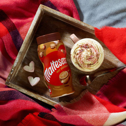 Maltesers hot chocolate mix and a cup of hot chocolate on a wooden tray with a red blanket underneath.