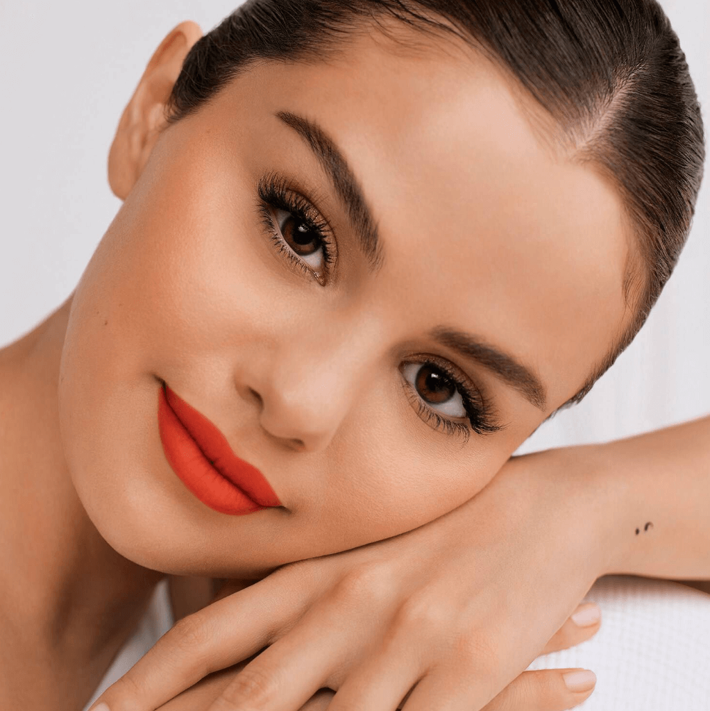 Model showcasing RARE BEAUTY Lip Soufflé Matte Lip Cream in vibrant orange shade, highlighting velvety texture and perfect color payoff.