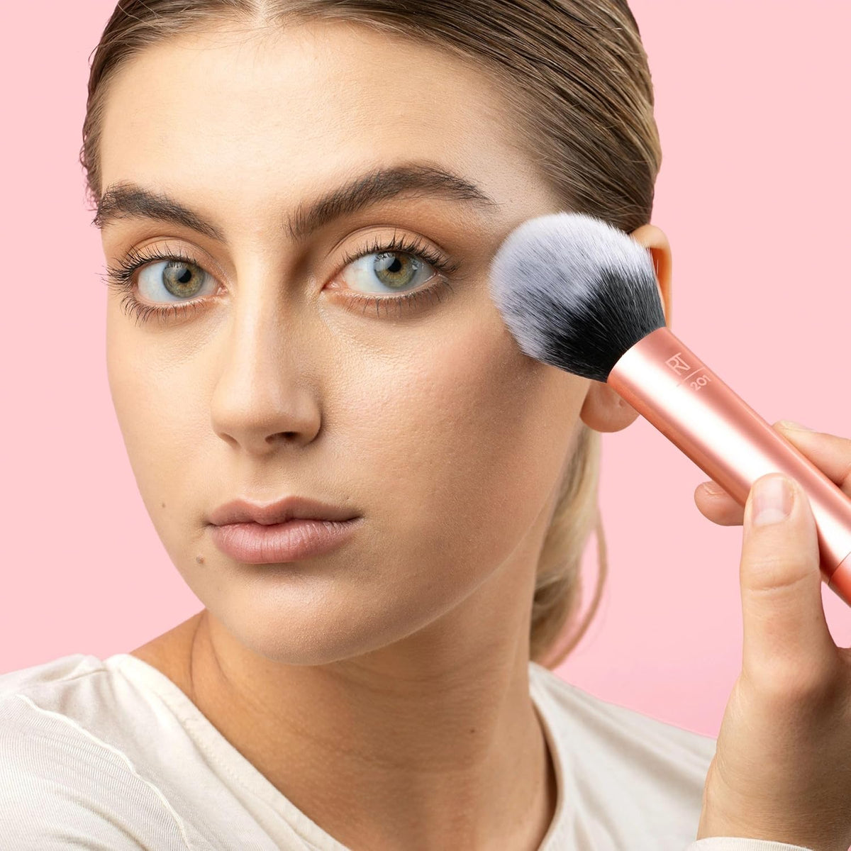 Woman applying makeup with a brush on a pink background