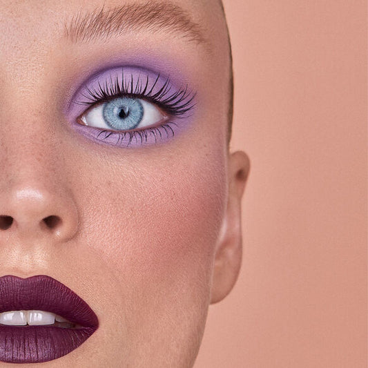 Close-up of a person's face with purple SWEED eye makeup and dark lipstick on a peach background