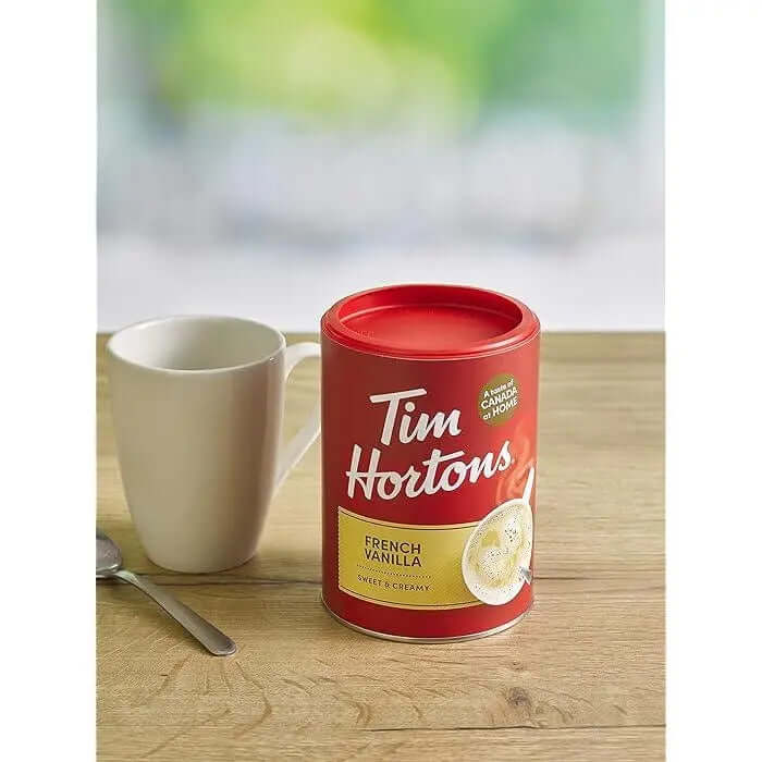 Tim Hortons Instant Cappuccino French Vanilla 454g can beside a white mug on a wooden table.