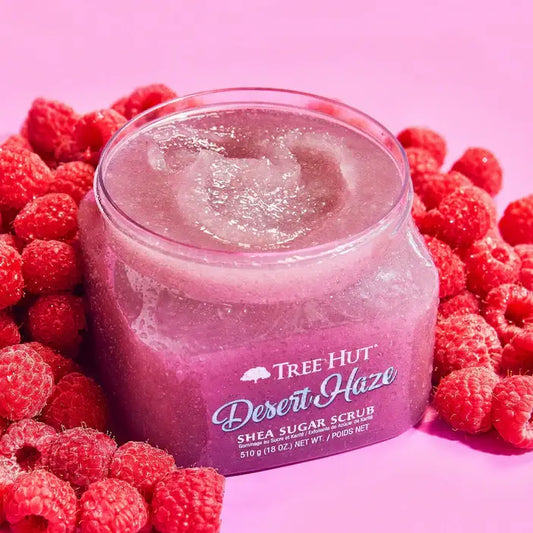 Jar of Tree Hut Desert Haze Shea Sugar Scrub surrounded by raspberries on a pink background