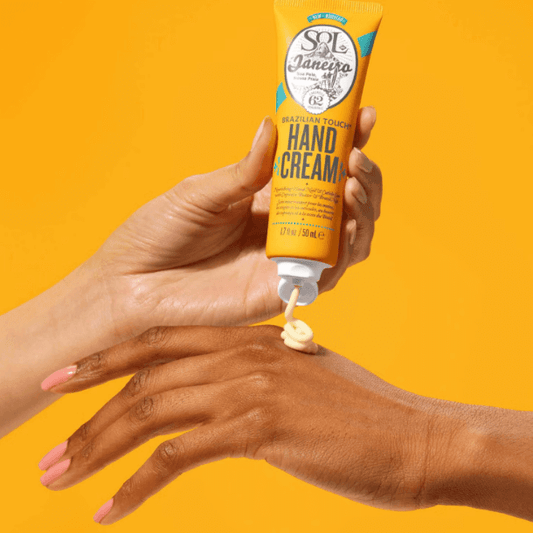 Sol de Janeiro Brazilian Touch Hand Cream 50ml being applied to hands with vibrant yellow background.