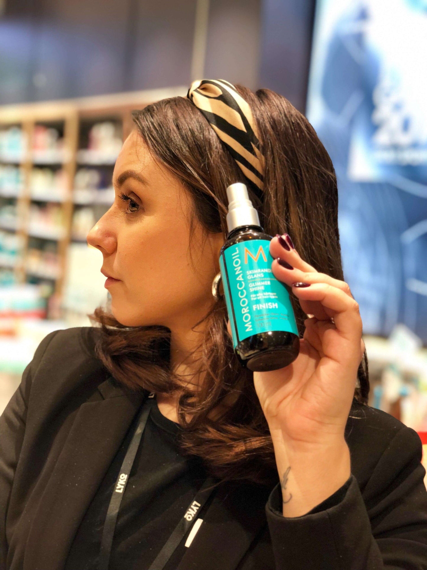 Woman showcasing Moroccanoil Glimmer Shine Spray 100ml in a beauty store, highlighting its shine-enhancing benefits.