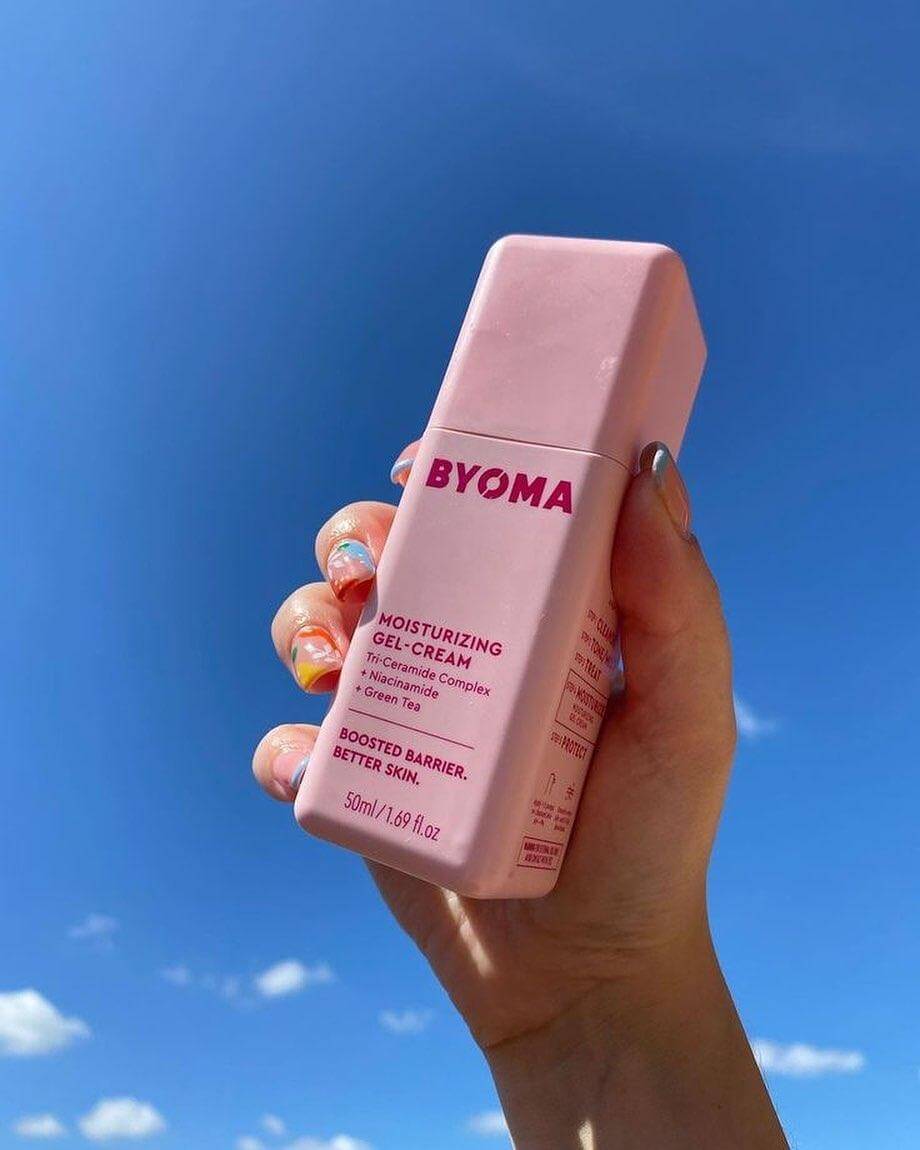 Hand holding BYOMA Moisturising Gel Cream 50ml against a blue sky, showcasing its colorful packaging.