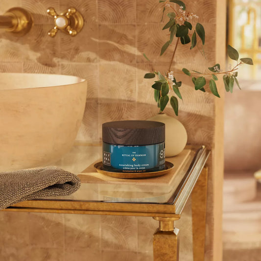 Jar of body cream on a wooden shelf in a bathroom setting with a plant and towel.