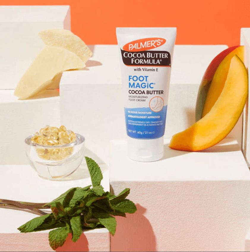 Palmer's Cocoa Butter Formula Foot Magic 60g cream with Cocoa Butter and Peppermint Oil on display with mango and mint.