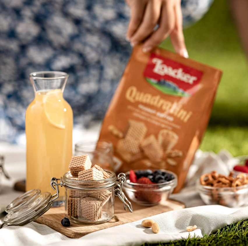 Enjoying Loacker Quadratini wafers with fresh fruits and a drink on a picnic blanket in the sun.