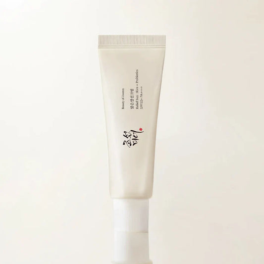 A single cosmetic cream tube standing upright against a plain off-white background. The tube is a warm off-white/cream color with black text and a small red accent, and has a white cap.