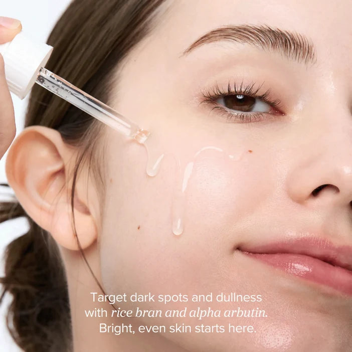 Close-up of a woman's face with a dropper applying a skincare product, text about rice bran and alpha arbutin.