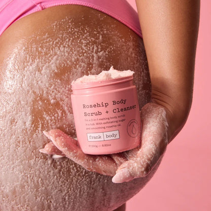 A close-up of a person's bare torso and hand covered in a white exfoliating body scrub, holding a pink jar labeled 'Rosehip Body Scrub + Cleanser' by frank body against a solid pink background.