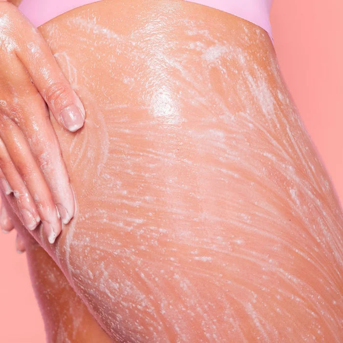 Close-up of a person's leg with exfoliating powder and a hand applying it against a pink background
