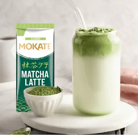 Mokate Classic Matcha Latte package next to a glass of creamy matcha latte and bowl of matcha powder on a light background.