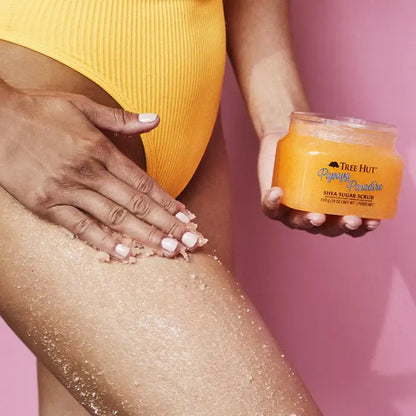 TREE HUT Sugar Scrub Papaya Paradise being applied to smooth skin, with bright yellow swimsuit against a pink background.