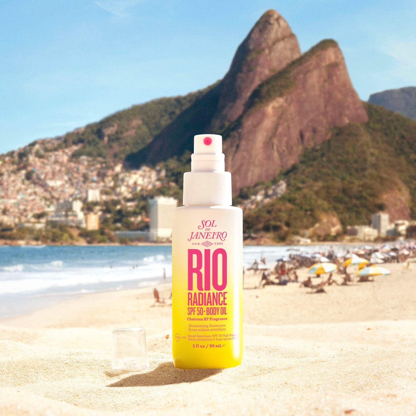 Sol de Janeiro Rio Radiance Oil SPF 50 90ml on a sandy beach with mountains in the background, perfect for sun protection.