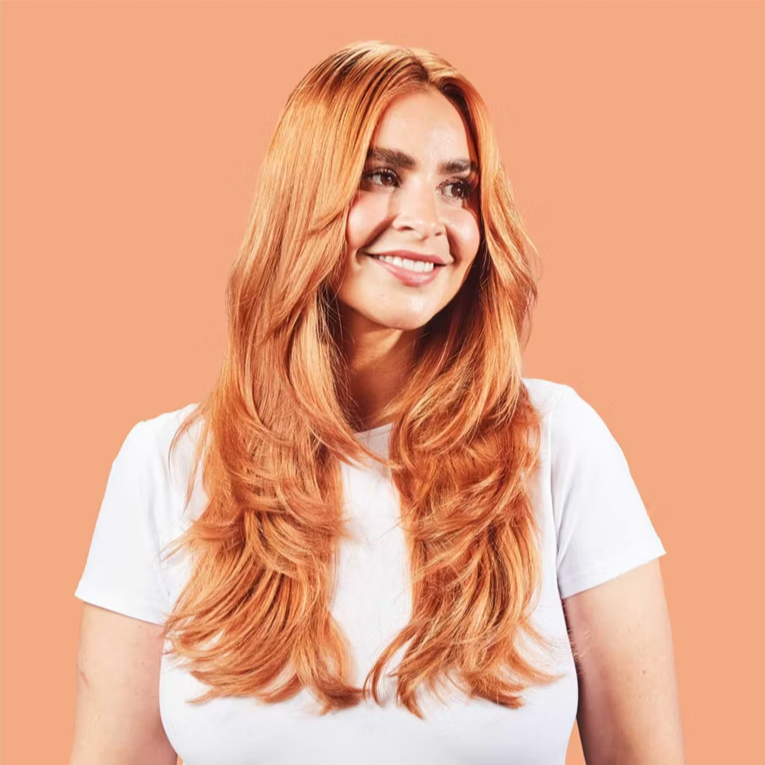 Woman with long, wavy red hair wearing a white t-shirt against a peach background