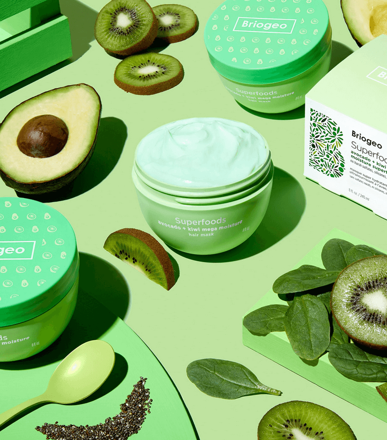 Briogeo skincare products with avocados, kiwis, and green leaves on a green background