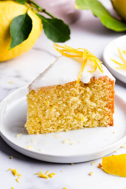 A slice of moist lemon cake topped with icing and lemon zest on a white plate, surrounded by fresh lemons.