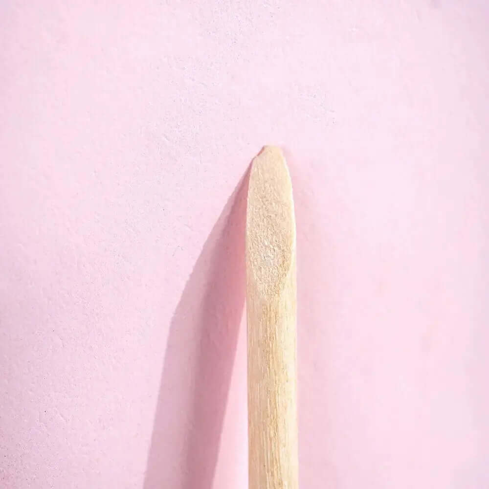 Close-up of a wooden cuticle stick against a pink background, showcasing its flat end for cuticle grooming.