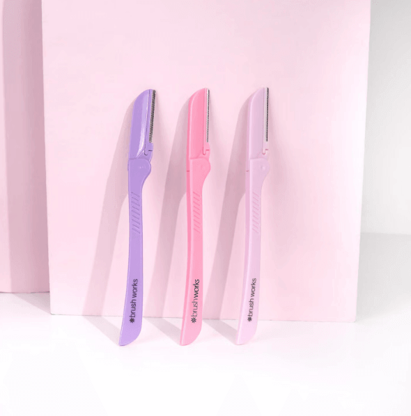 Brushworks Eyebrow Razors in purple and pink for pain-free brow shaping and exfoliation on a pink background.