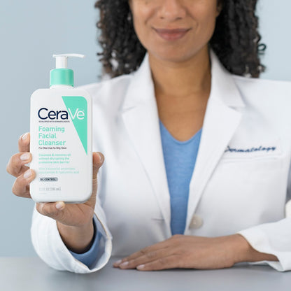 Person in a white coat holding a CeraVe foaming facial cleanser bottle against a light gray background