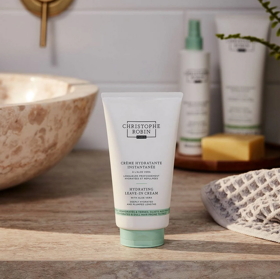 Tube of Christophe Robin hydrating hair cream on a bathroom shelf with other products.