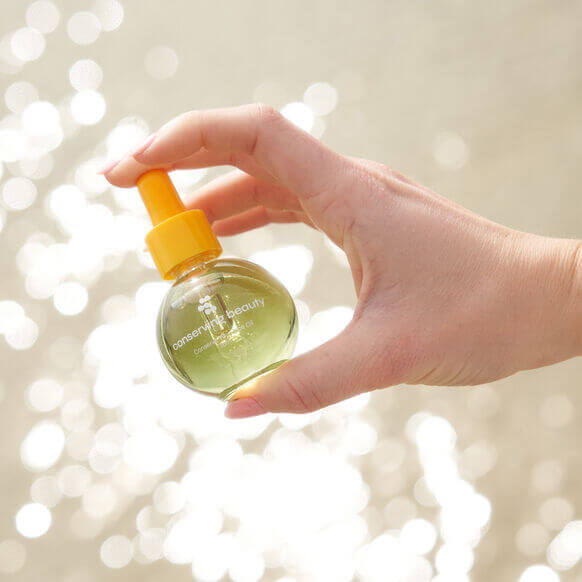 Hand holding Conserving Beauty Conserve You Face Oil bottle with a shimmering background, highlighting its radiant skin benefits.