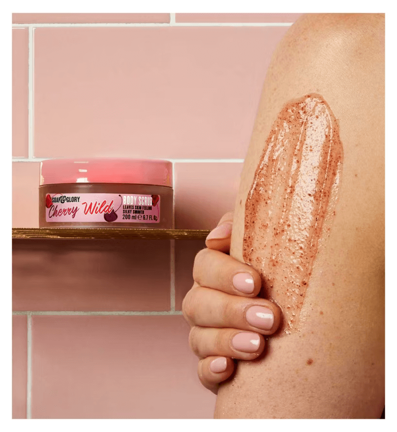 Close-up of Soap & Glory Cherry Wild Body Scrub being applied on a woman's arm in a pink tiled bathroom.