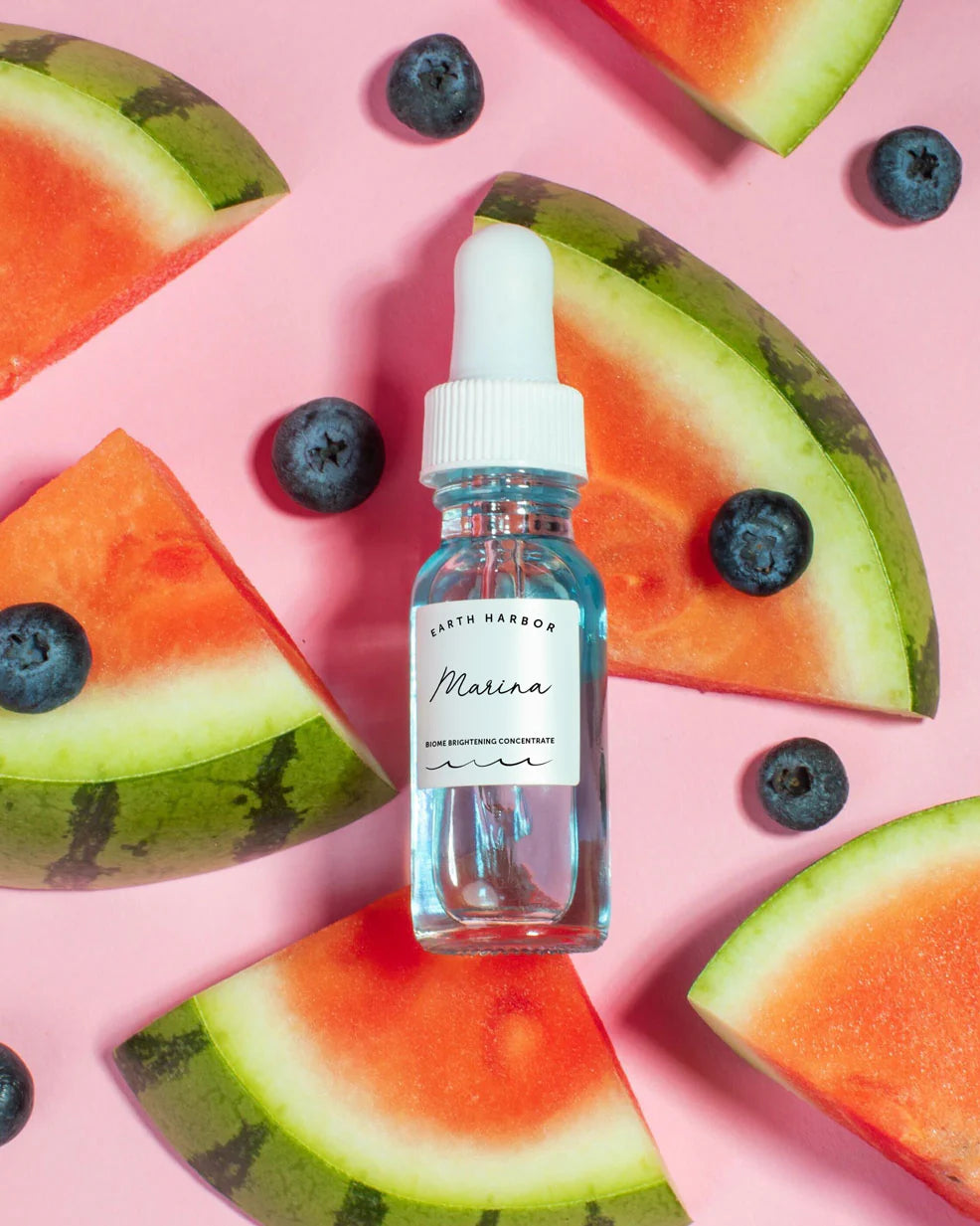 Earth Harbor Bottle of Marina skincare product surrounded by watermelon slices and blueberries on a pink background