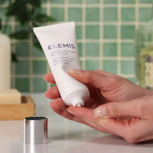 Hand holding an Elemis skincare product against a green tiled wall.