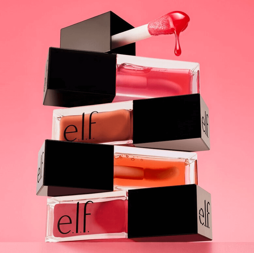 Stack of e.l.f Glow Reviver Lip Oil bottles with a glossy red applicator against a pink background.