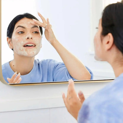 Woman applying First Aid Beauty Face Cleanser while looking in the mirror, promoting healthy skin routine.