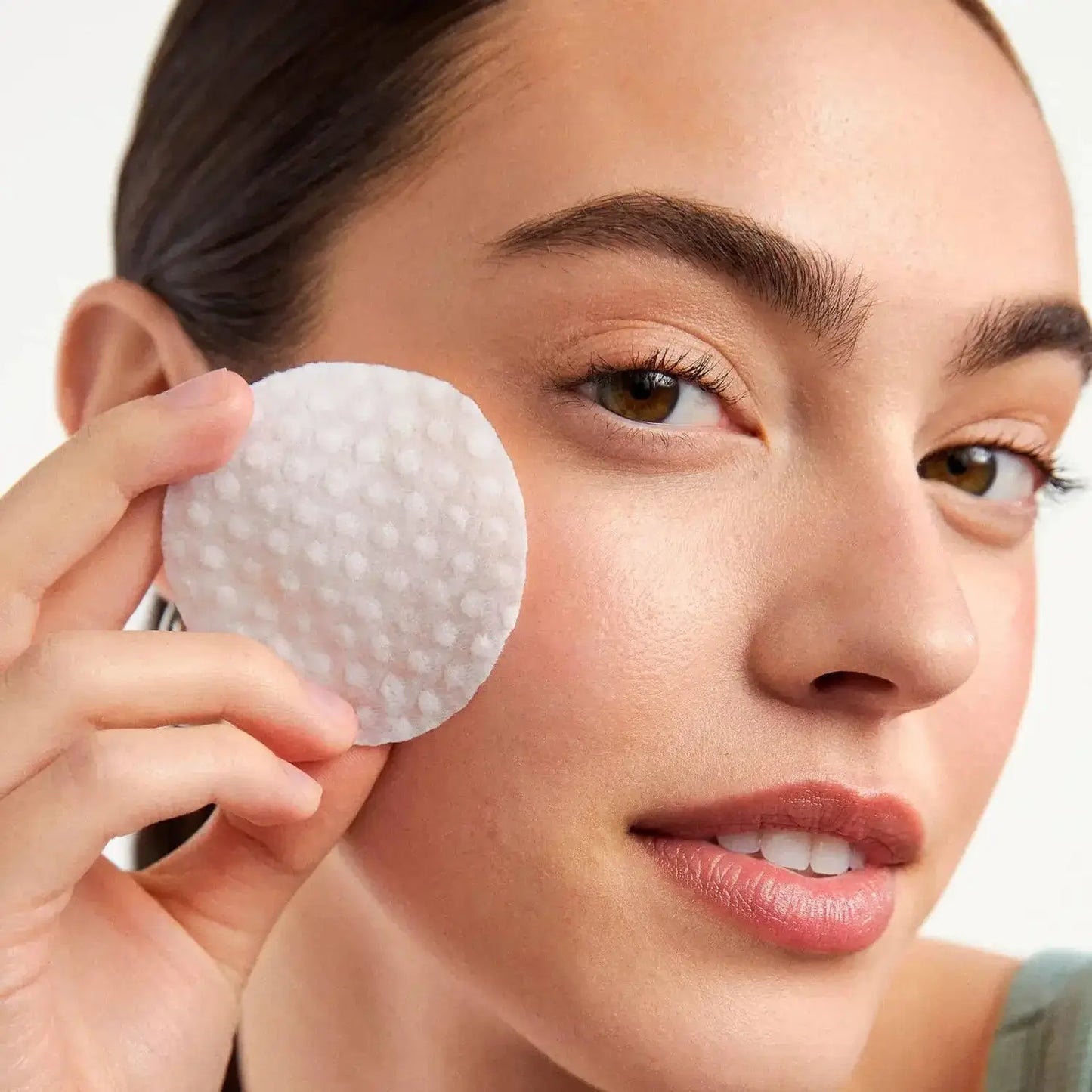 A woman holding a First Aid Beauty Facial Radiance Pad to her cheek, showcasing gentle exfoliation for brighter skin.