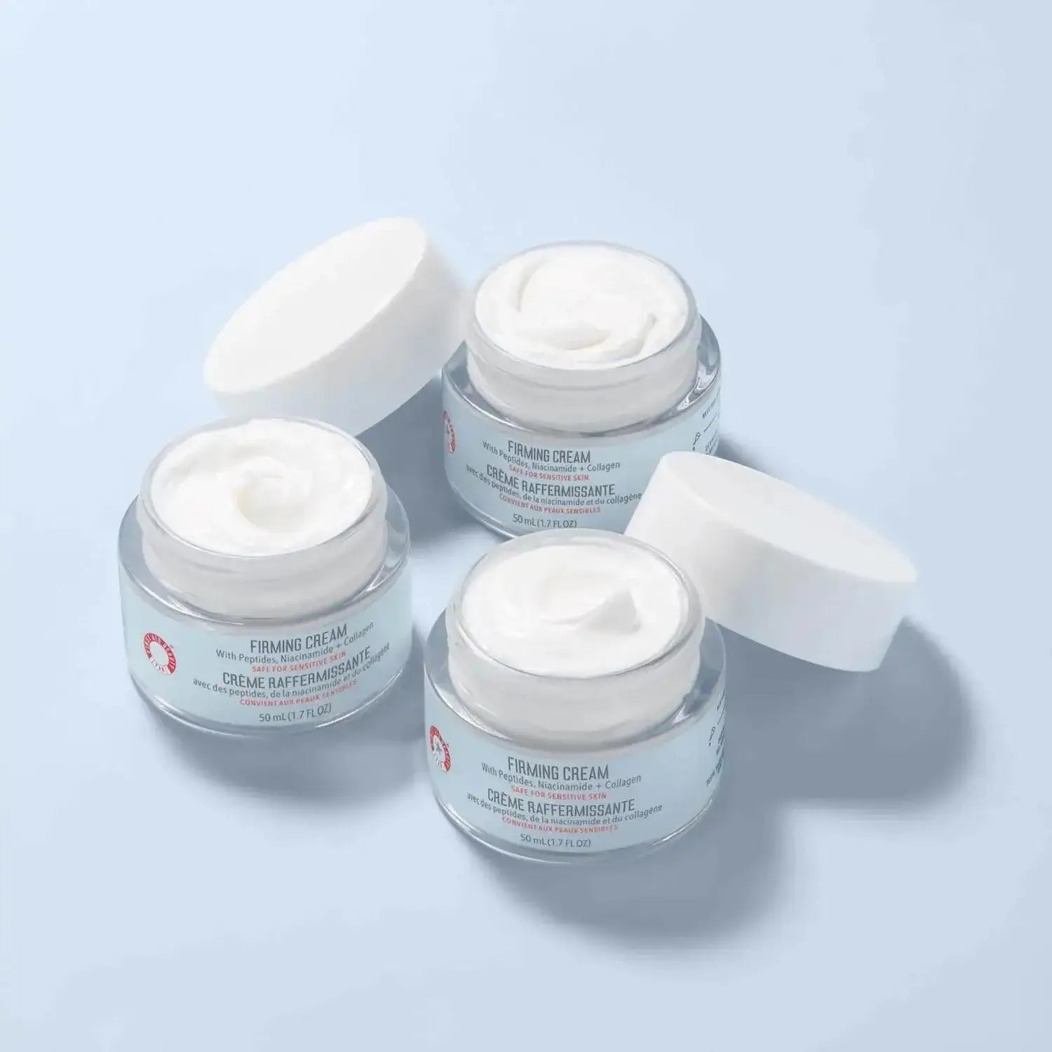 Three jars of First Aid Beauty Firming Cream with Peptides, Niacinamide + Collagen on light blue background.