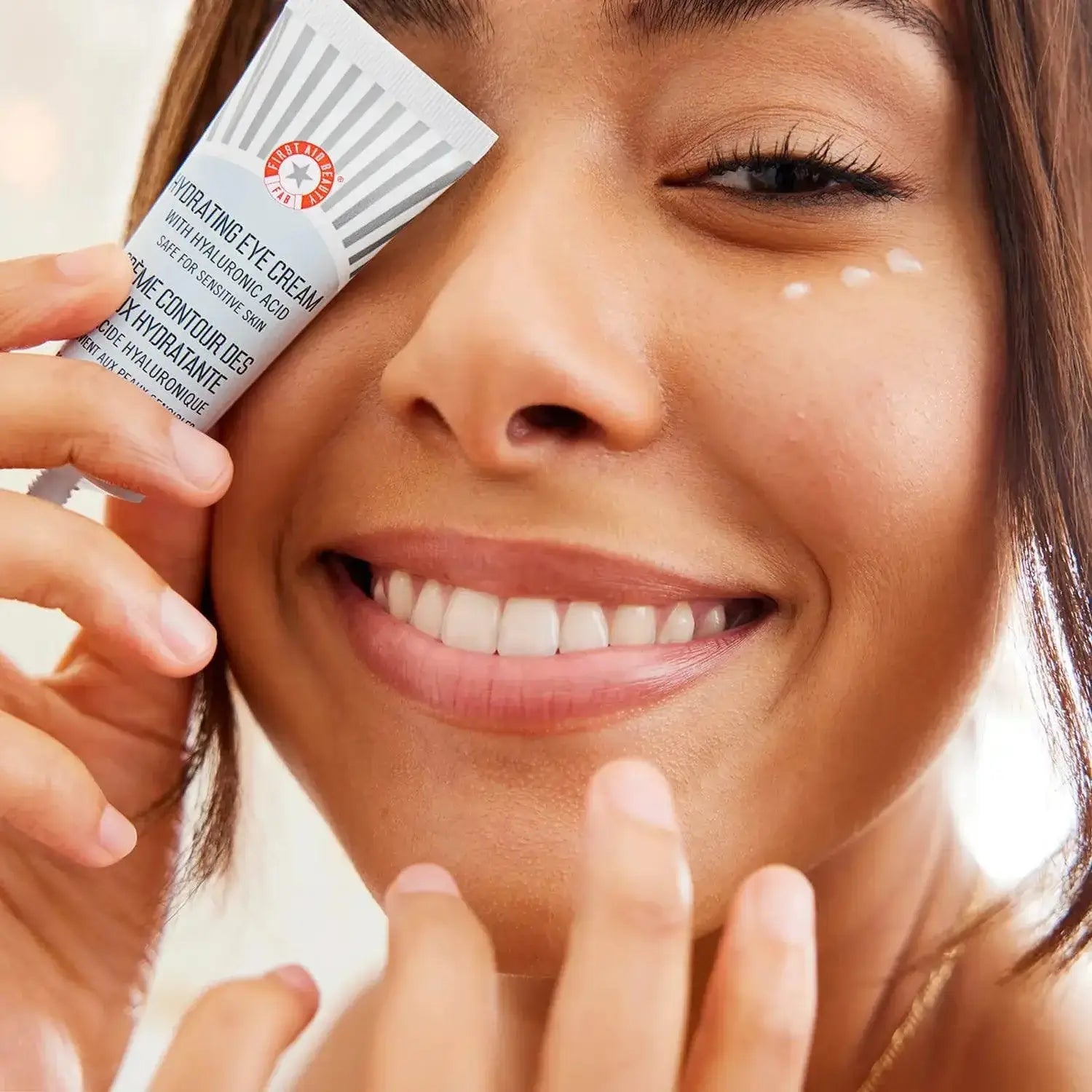 Smiling woman applying First Aid Beauty Hydrating Eye Cream 15ml to her eye area for moisture and brightness.