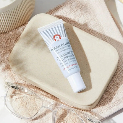 First Aid Beauty Hydrating Eye Cream 15ml tube on a beige plate, designed to moisturize and reduce puffiness.