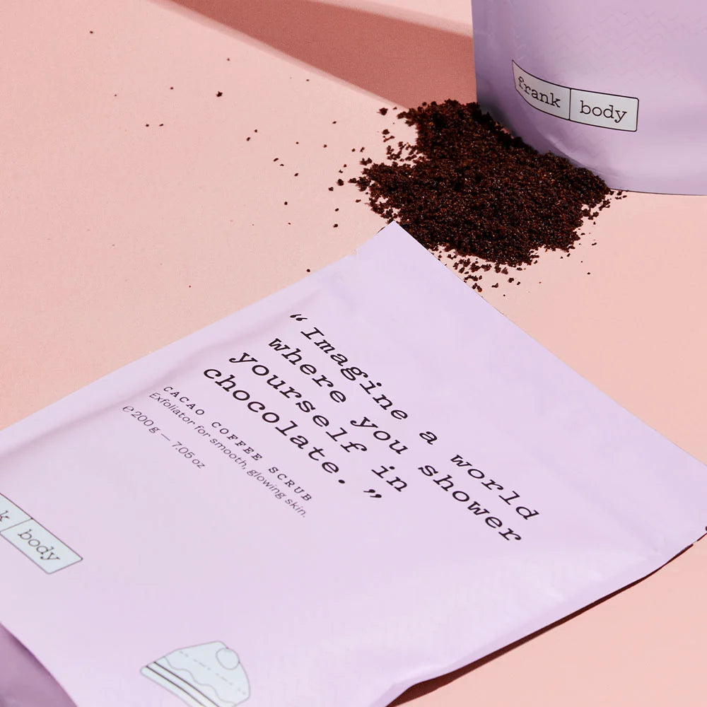 A light lavender sachet of Frank Body cacao coffee scrub lies on a pink surface, with dark brown scrub spilled beside it and part of the product container visible in the background.