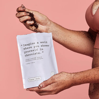 Person holding a cacao coffee scrub packet with text and 'frank body' branding on a pink background.